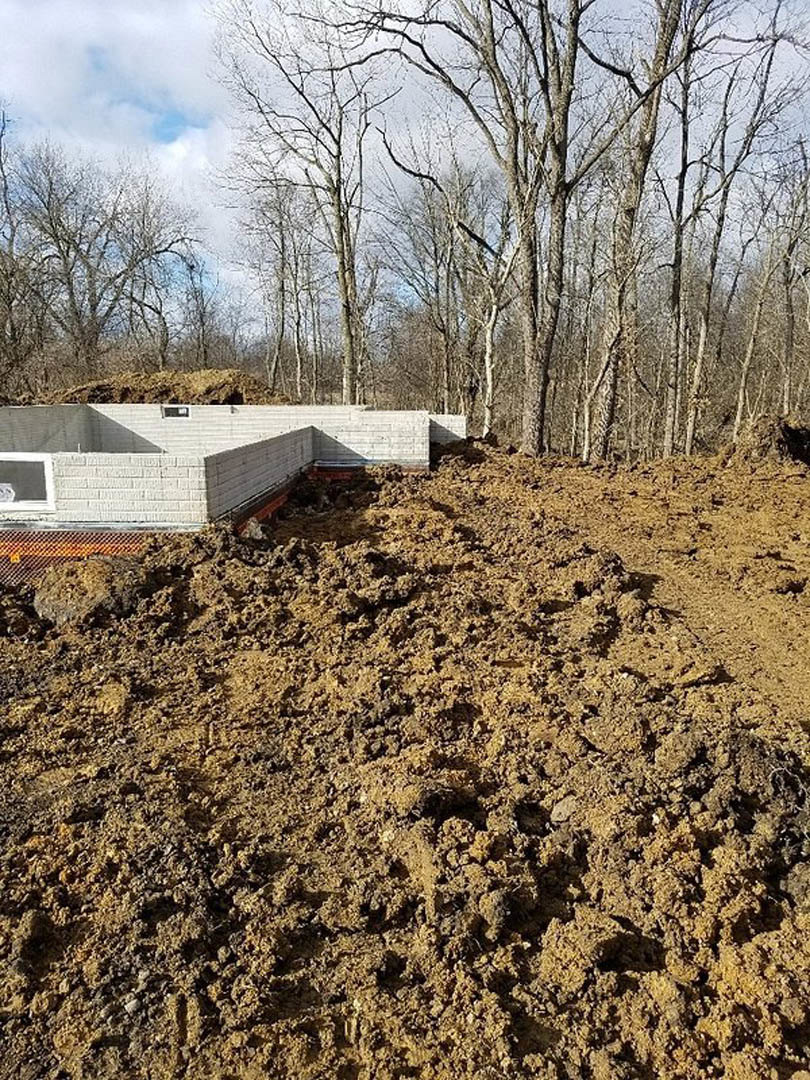 Freshly excavated soil and dirt piles beside a partially constructed building with bare trees and cloudy sky in the background