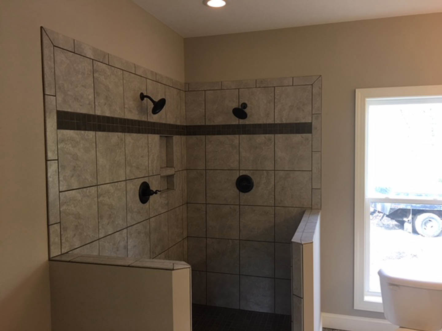 Modern bathroom with a glass-enclosed shower featuring a tiled wall, white toilet, wall-mounted mirror, and window overlooking a trailer outside