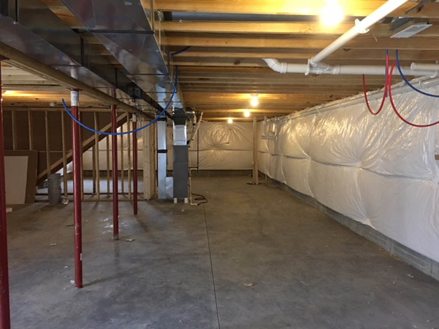 Basement with concrete floor, white plastic sheeting covering walls, exposed ceiling beams, and a red object near the wall