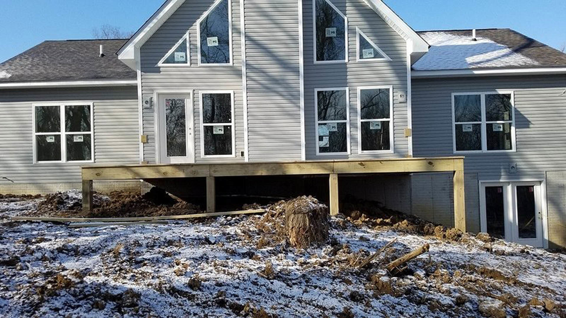 Two-story house under construction with gray siding, double front doors, white-framed windows displaying permit signs, snow-covered ground with rocks and dirt, and a pile of soil