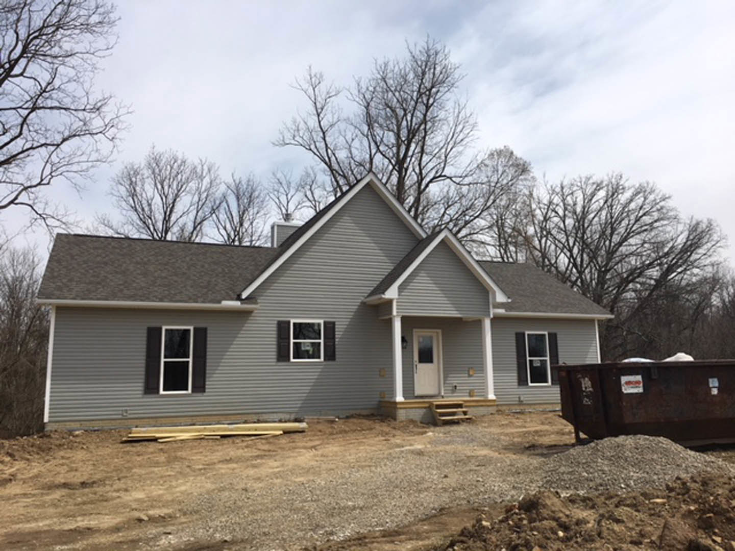 Partially built house surrounded by dirt and mature trees, white framed windows and door visible, metal container with white sign nearby, cloudy sky overhead