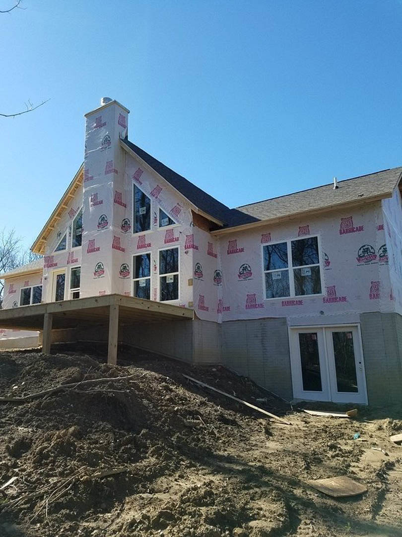 Two-story house under construction with exposed framing, glass double doors, large windows, and a pile of dirt in the foreground