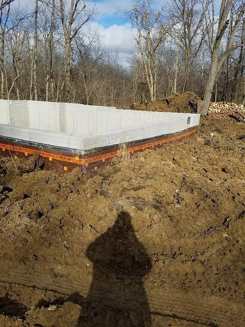 Shadow of a person cast on a dirt surface near a large white box with orange and black stripes, surrounded by trees and plants under a partly cloudy sky