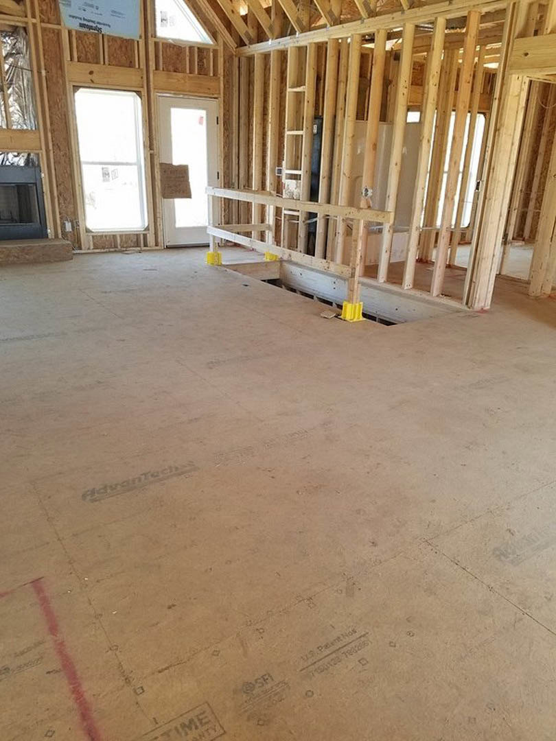 Unfinished room with exposed wood framing, plywood floor, wall opening, black-framed window, and paper taped to a doorway
