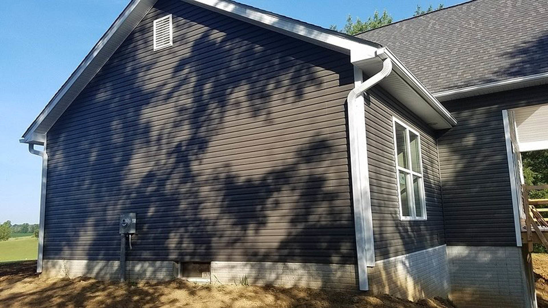 Two-story house with light siding, white gutters, shingle roof, and large windows; wooden fence in foreground, blue sky above