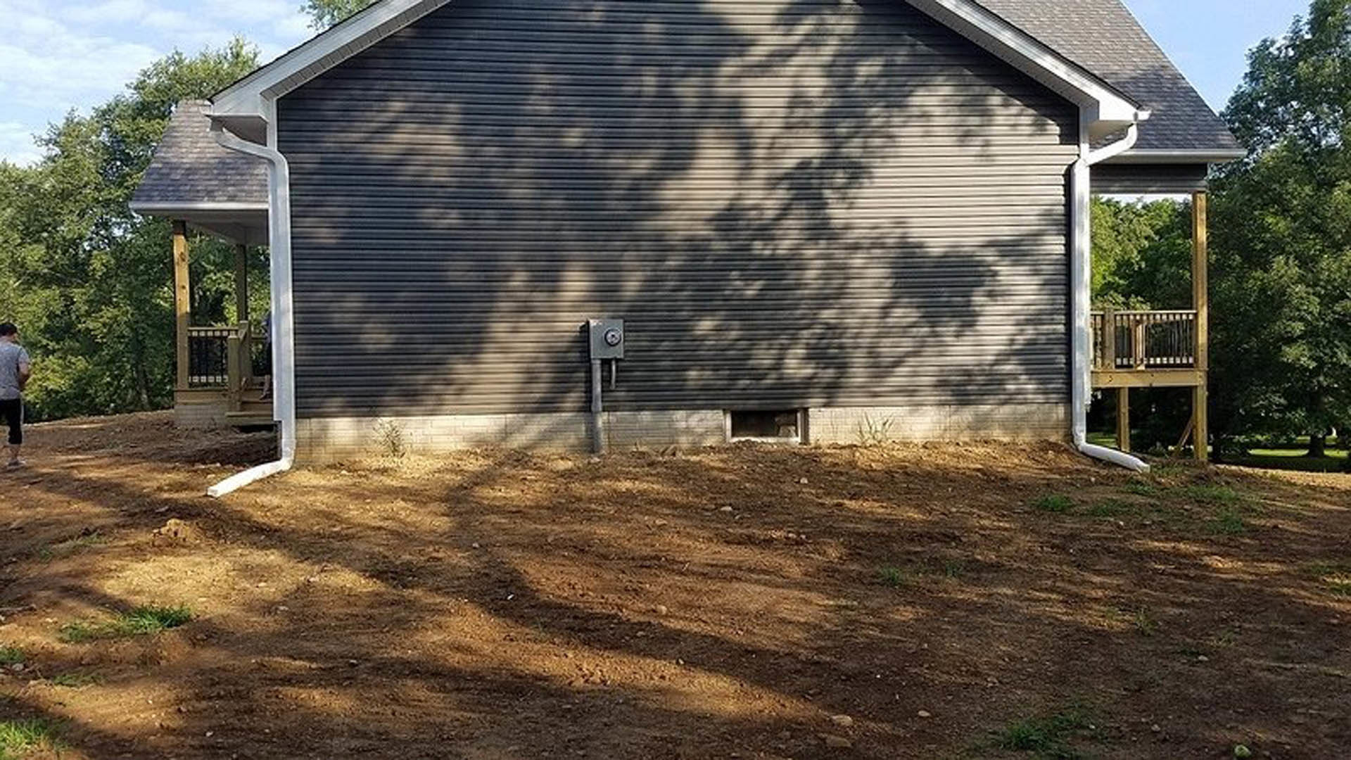 Single-story cottage with light siding, surrounded by a large bare dirt yard, mature trees, and a small shed in the background.