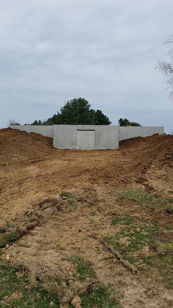 Modern home exterior with light gray siding, white door, unfinished landscaping, exposed soil, and surrounding trees under a partly cloudy sky