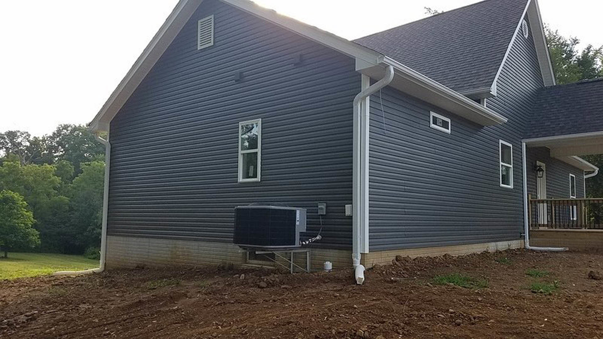 Two-story home with light siding, prominent outdoor air conditioning unit beside the foundation, large windows, and mature trees in the yard.