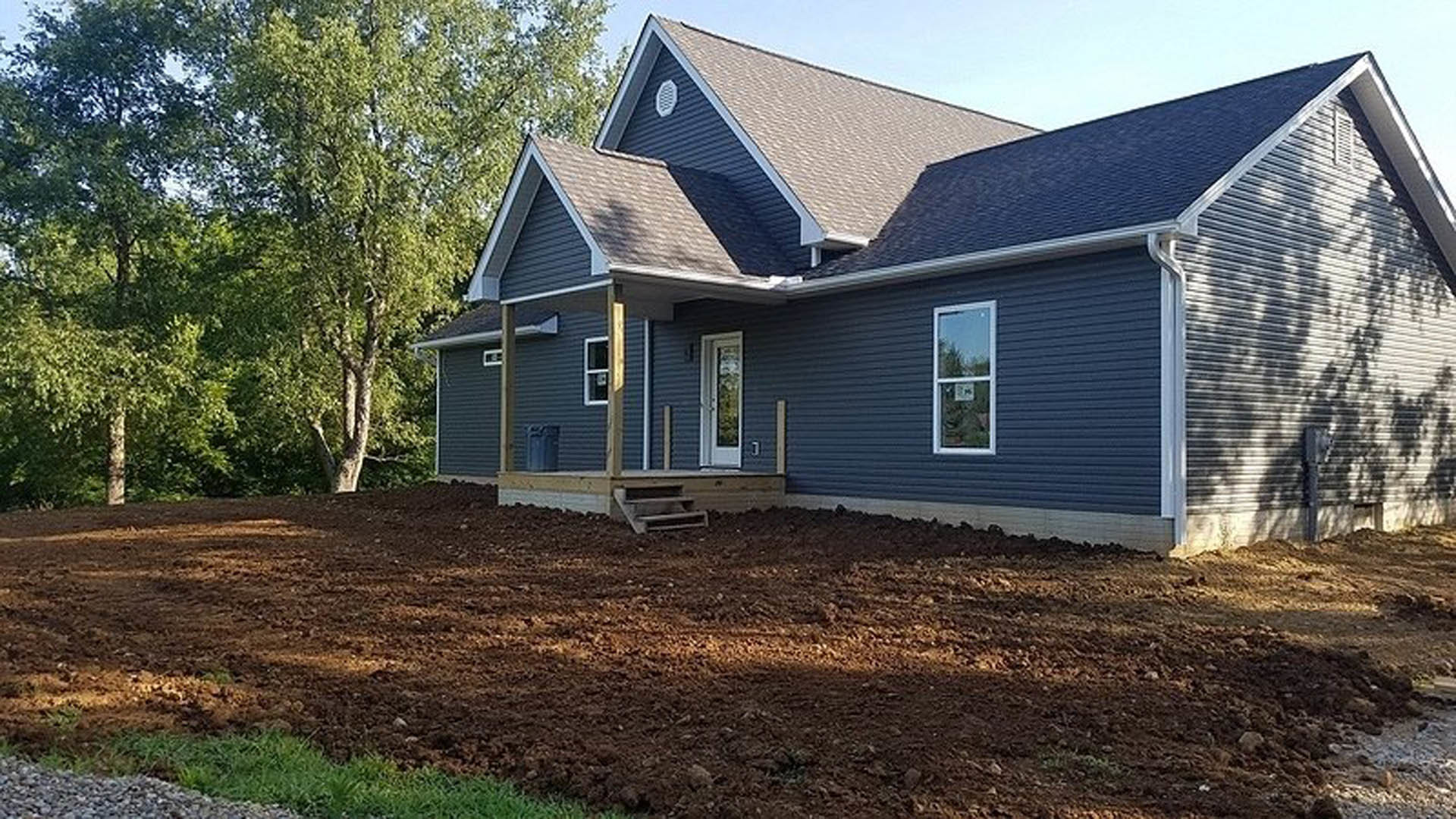 Two-story house with light-colored siding, large windows, and bare dirt yard in front, surrounded by trees under a clear sky