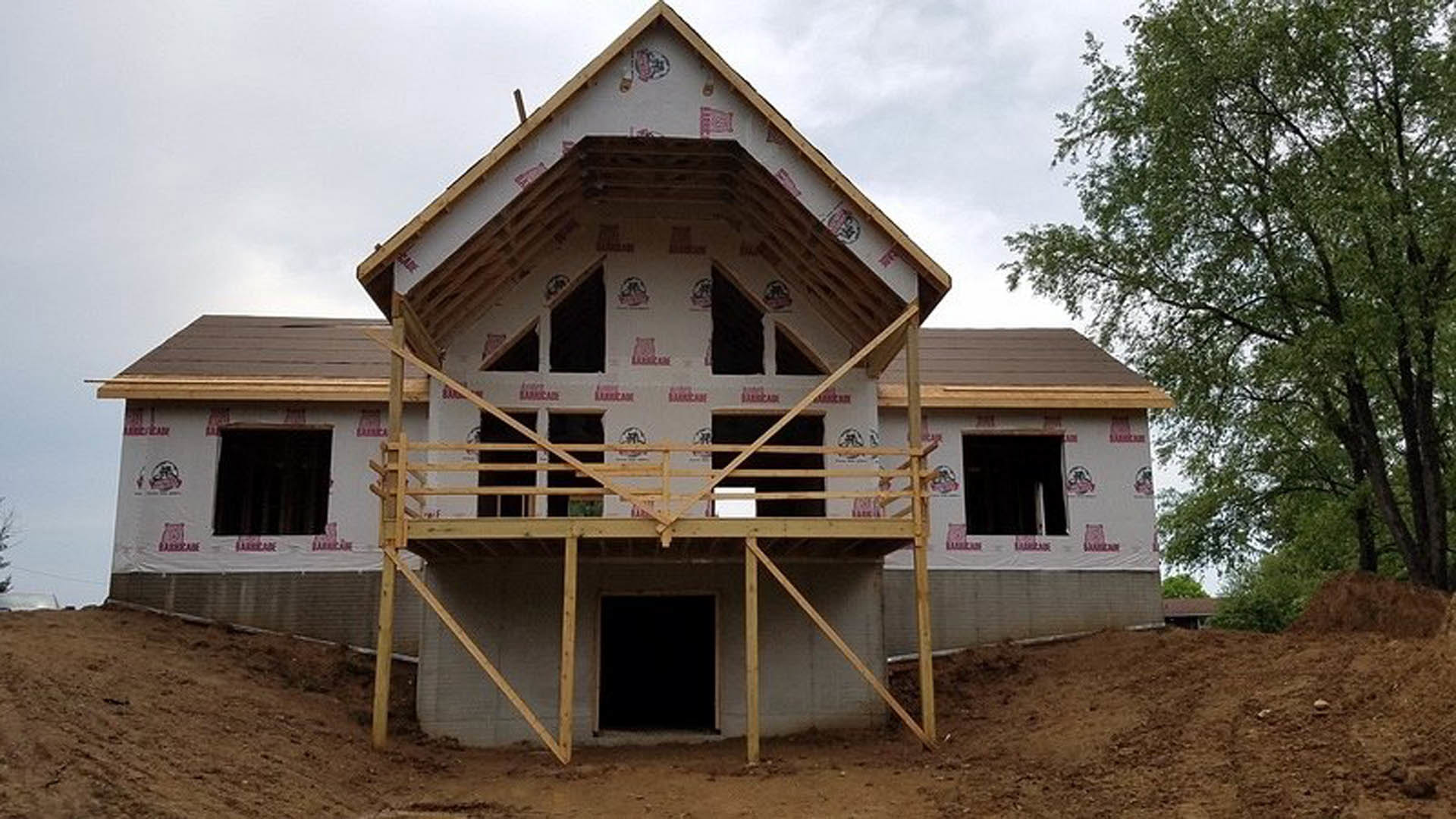 Two-story home under construction featuring a second-floor balcony with black-framed windows, exposed wooden framing, and a tree visible in the background.