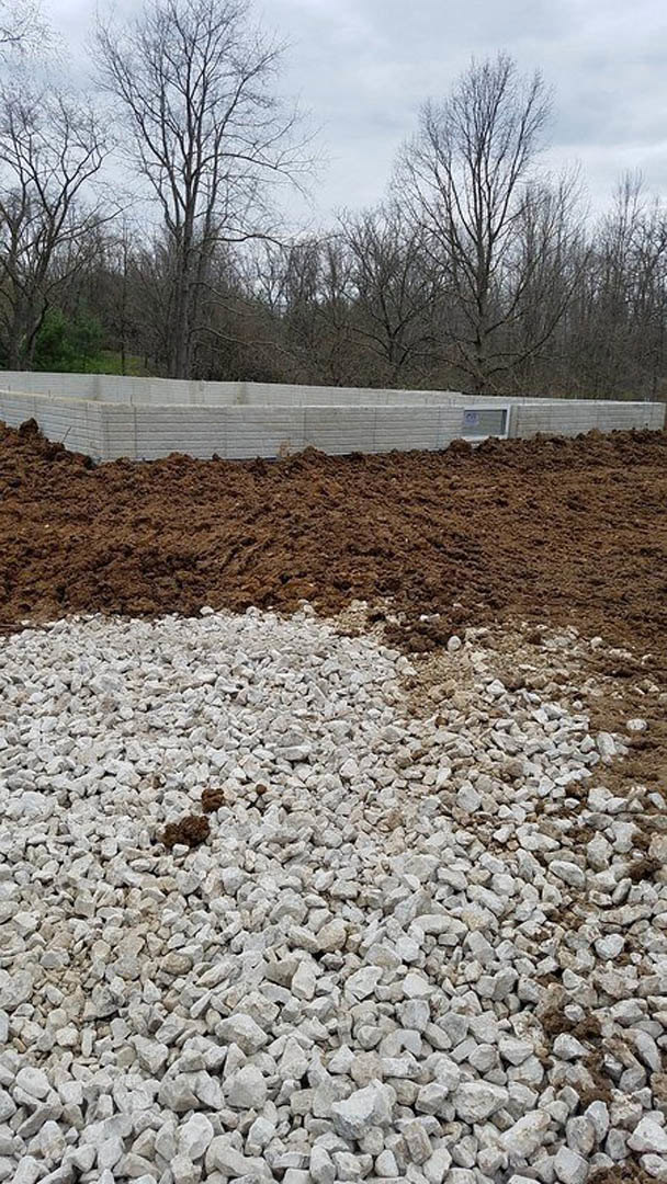 Pile of dirt and white gravel beside a white brick building, leafless tree and forested background, winter landscape