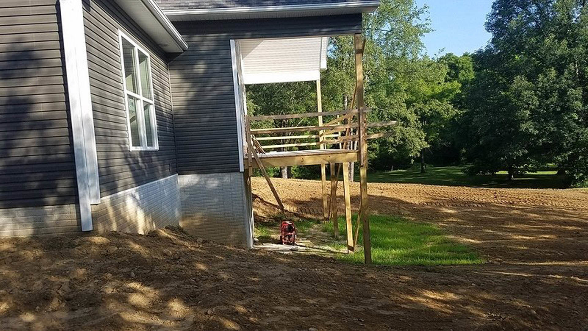 Wood-framed deck under construction attached to a light-colored house, red toolbox in the background, ladder leaning against siding, surrounded by trees and yard
