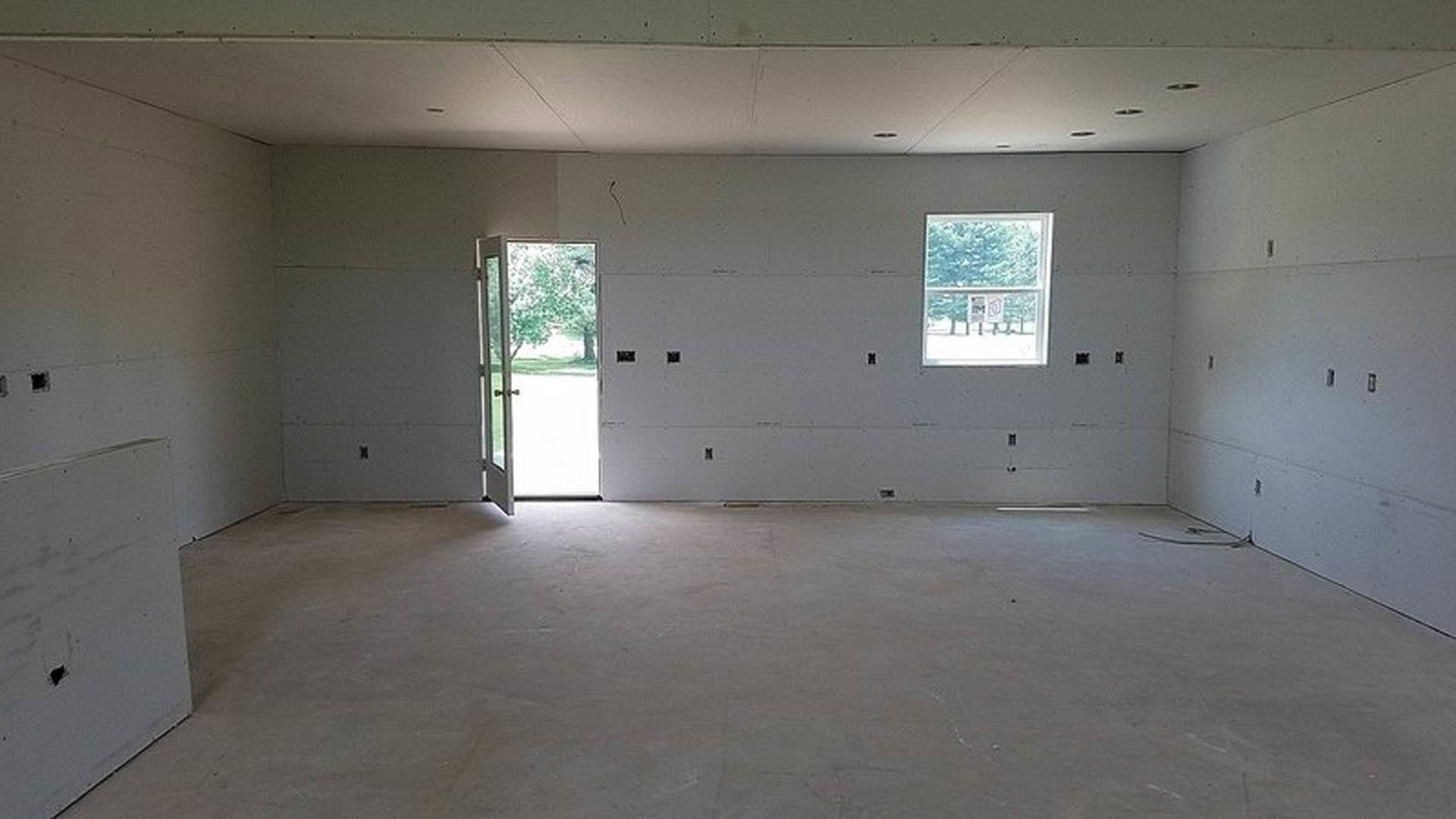 Sunlit room with light wood flooring, white plaster walls, open glass door, large windows, and tree visible outside