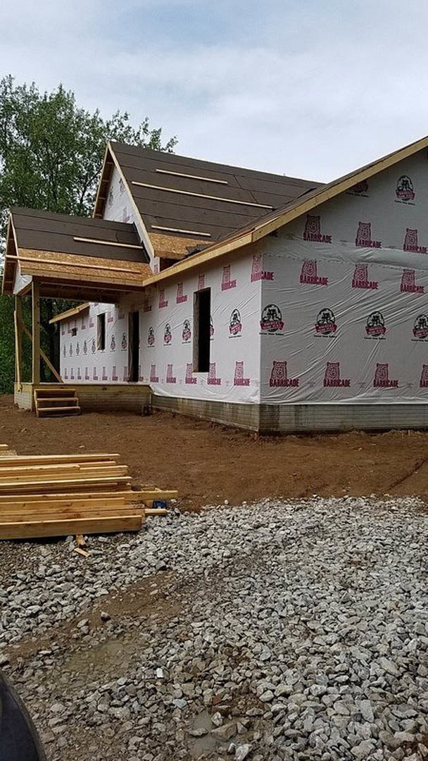 Partially built house with exposed wooden framing, white tarp with red logos covering sections, pile of wood planks and rocks on dirt ground, cloudy sky overhead