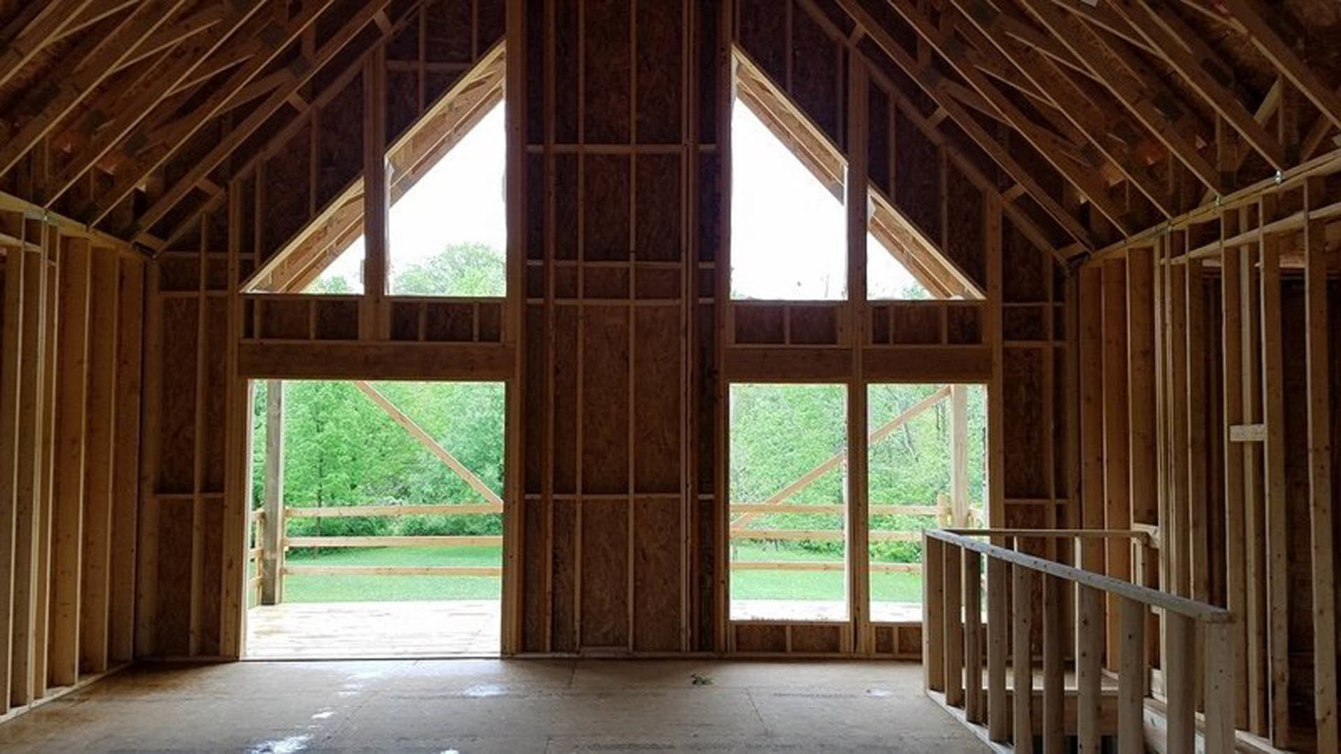 Wood-framed house under construction featuring a balcony with wooden railing, exposed beams, unfinished plank flooring, and large windows allowing daylight into the interior.