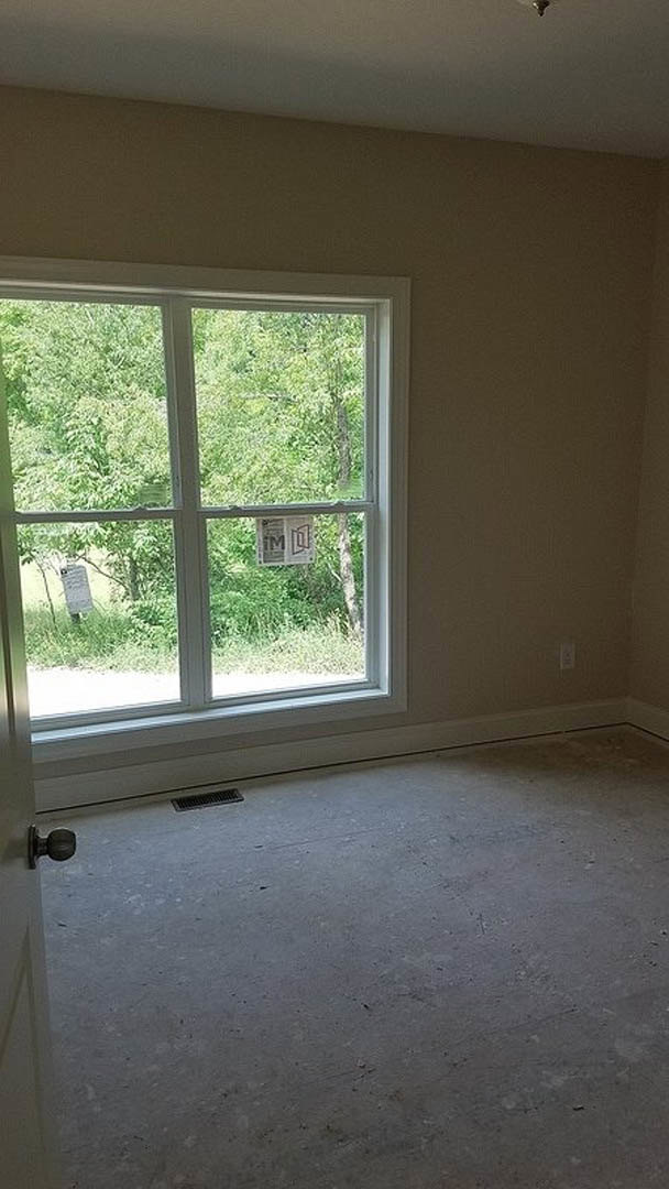 Bright room with large window framed by white trim, grey plank flooring with floor vent, trees visible outside, small sign attached to window glass