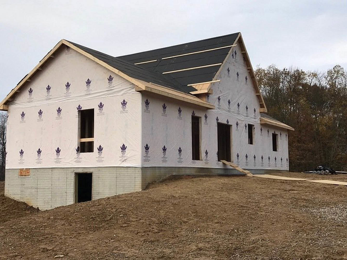 Partially built house with black roof, white walls, and black door, set against a dirt hill and surrounded by trees under a cloudy sky