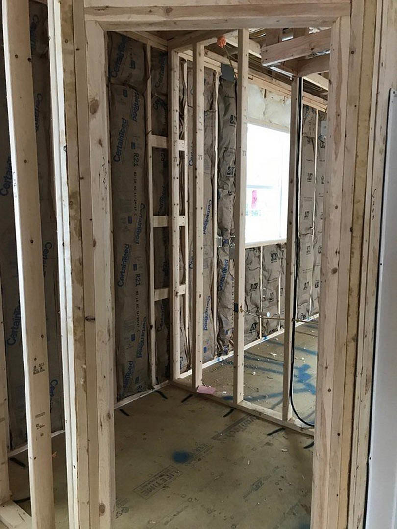 Wood-framed room under construction with exposed beams, white door featuring a pink sign, stained subfloor, window set in unfinished wall, and brown paper bag with blue writing on