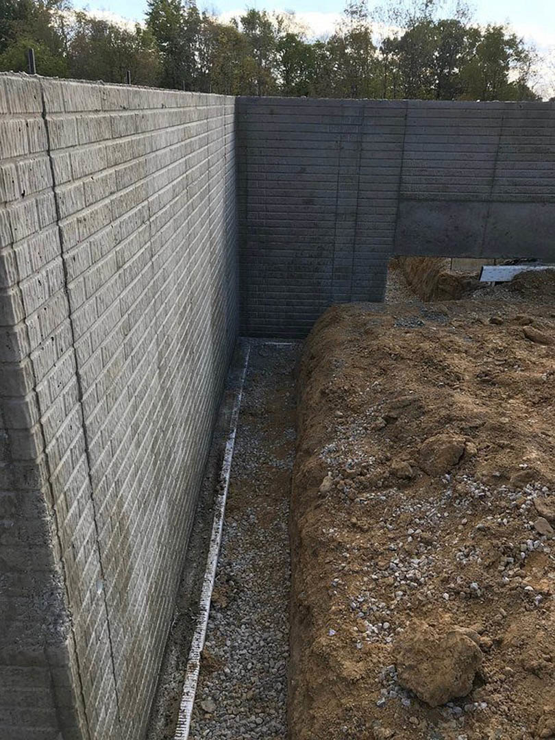 Concrete exterior wall with central hole, dirt ground in foreground, distant trees and sky visible outdoors