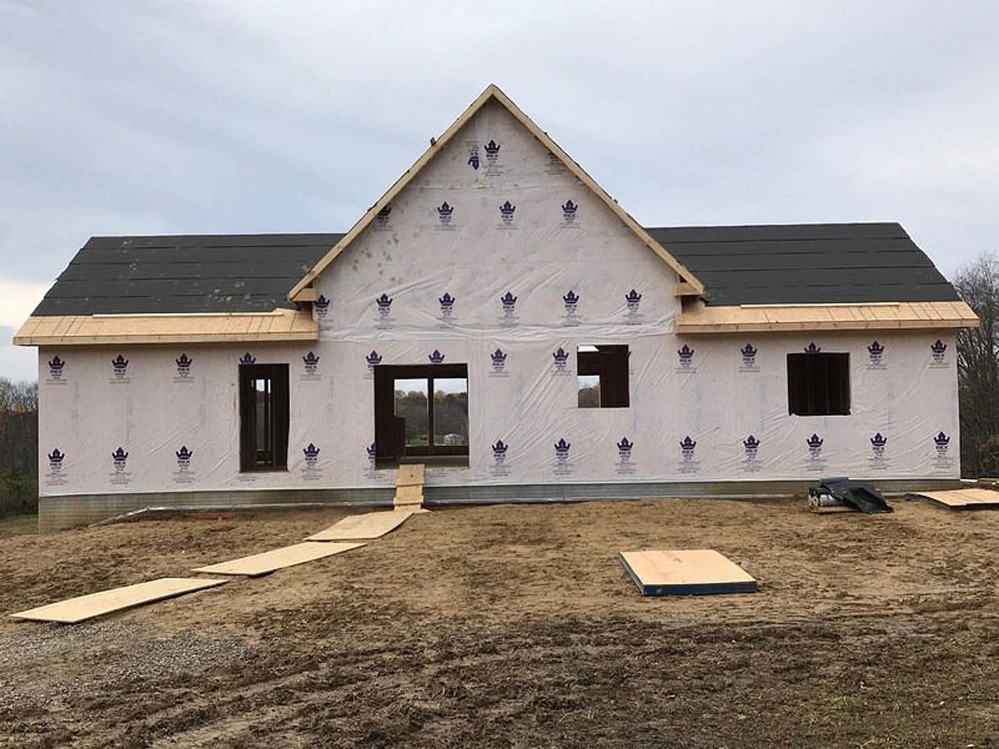 Two-story house under construction with plastic sheeting covering exterior walls, exposed dirt yard, large windows reflecting trees, and wooden boards scattered on ground.