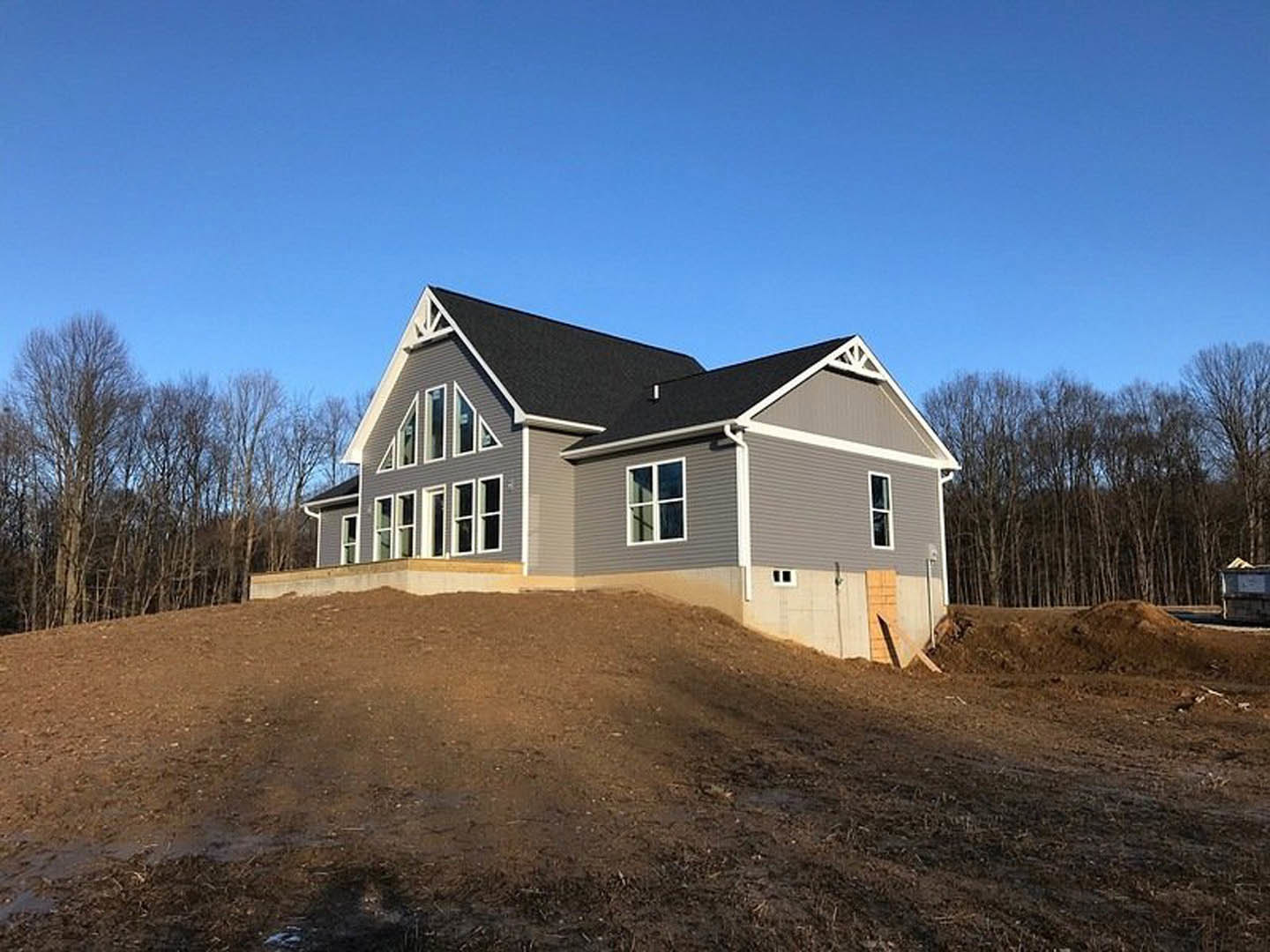 Two-story house with white-framed windows and light siding, situated atop a dirt hill with mature trees and blue sky in the background