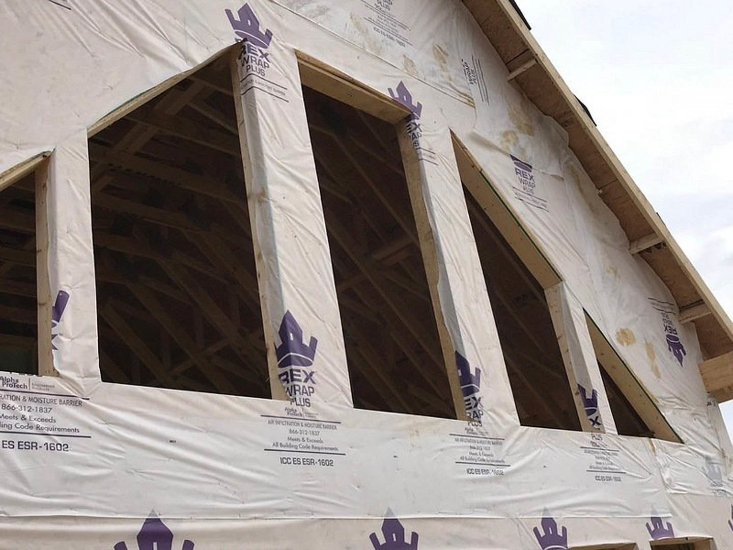 Wood-framed house under construction with white plastic sheeting covering exterior walls, exposed doorway, and visible windows under a clear sky.