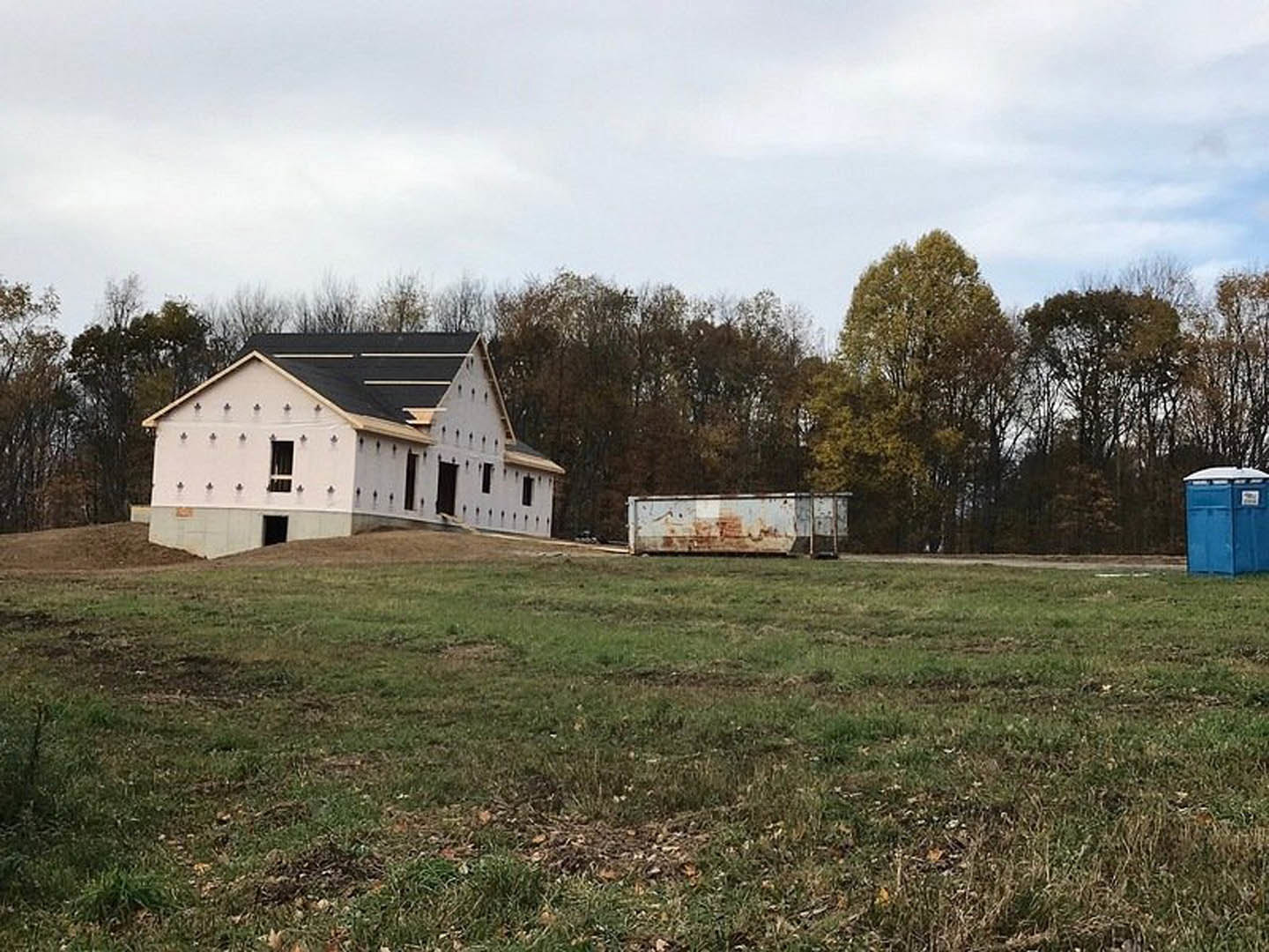 White house with black roof set on grassy lot, large white building in background, scattered trees and blue waste container nearby under cloudy sky