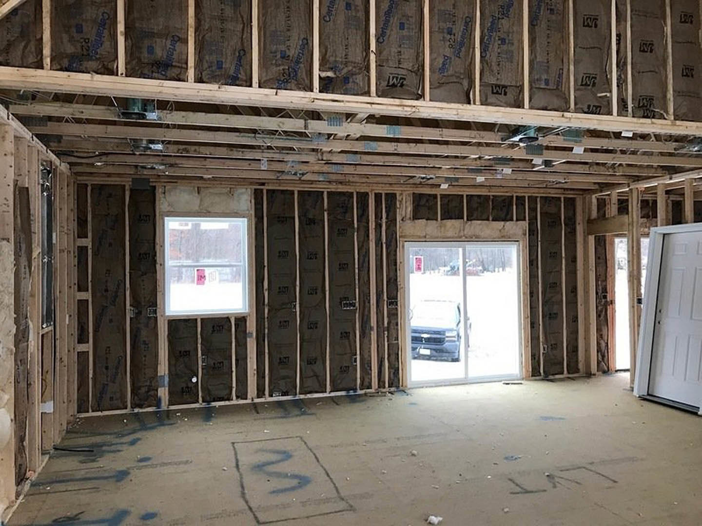 Open room with exposed wood ceiling beams, large windows, white door, concrete floor with black paint, parked car visible outside, pink sign on window