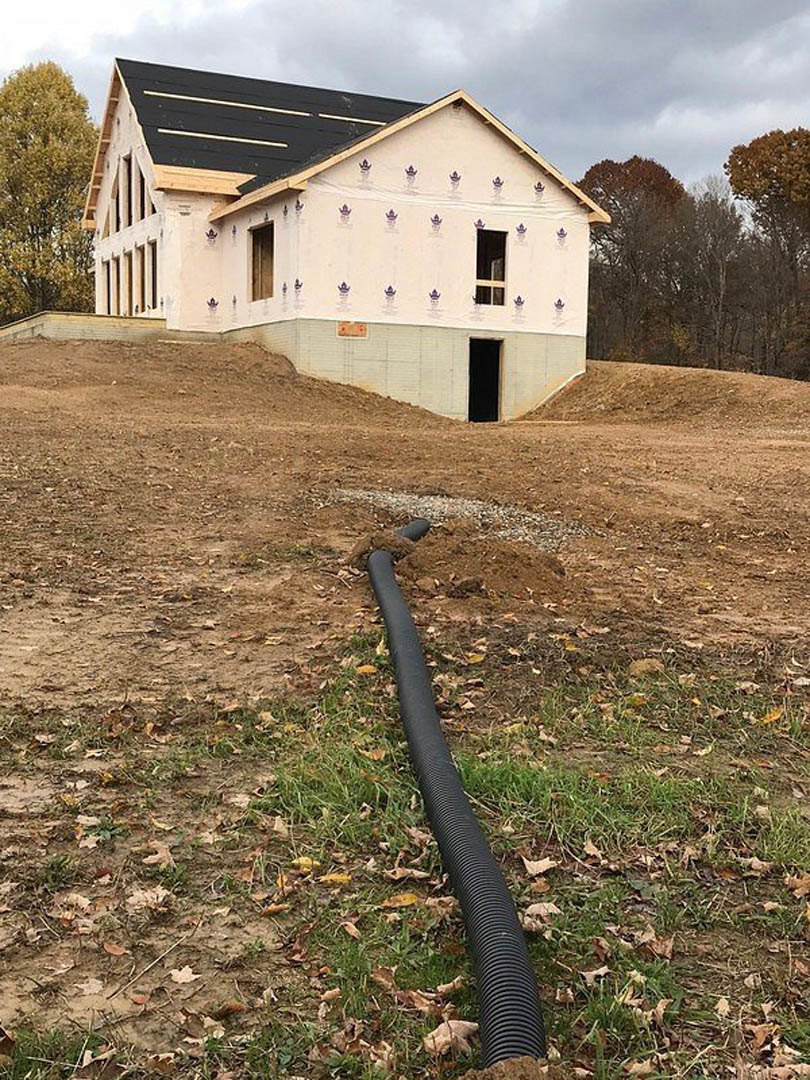 Black pipe lying in dirt in front of a partially constructed house with black roof and white stripes, surrounded by grassy land, green-leaved tree, and cloudy sky.