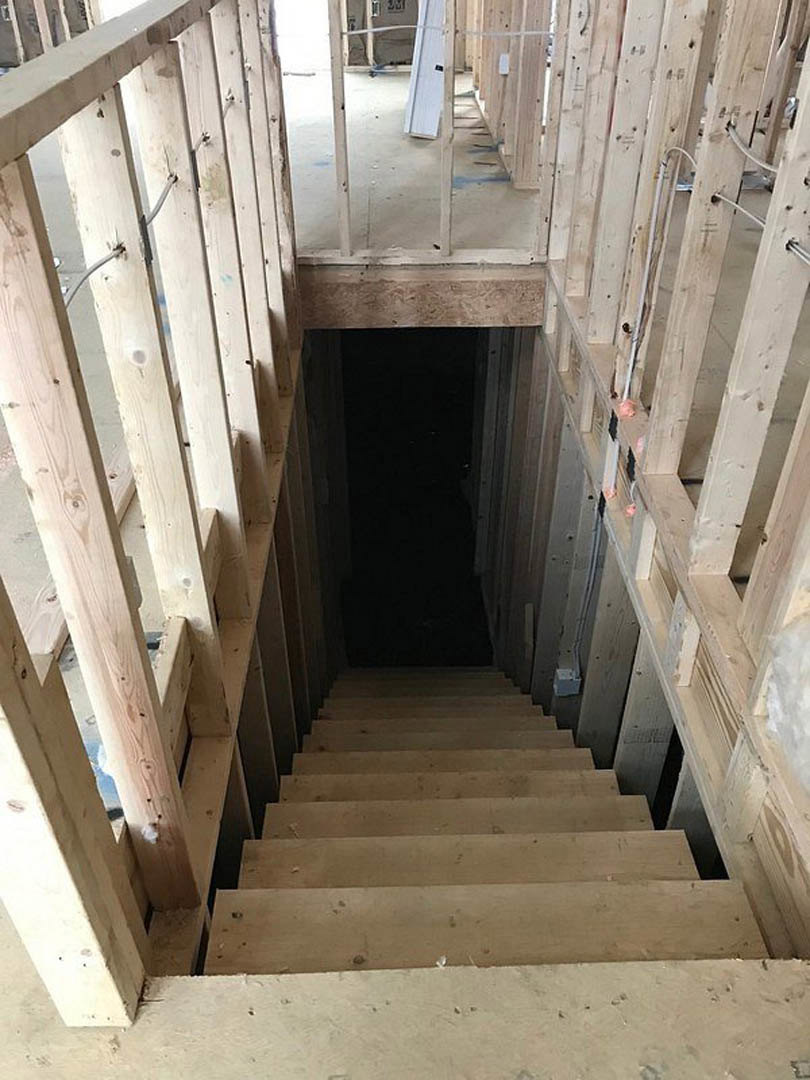 Wood staircase with white risers ascending through an opening in the ceiling, surrounded by dark walls, exposed wood beams, and a white door in a modern interior.