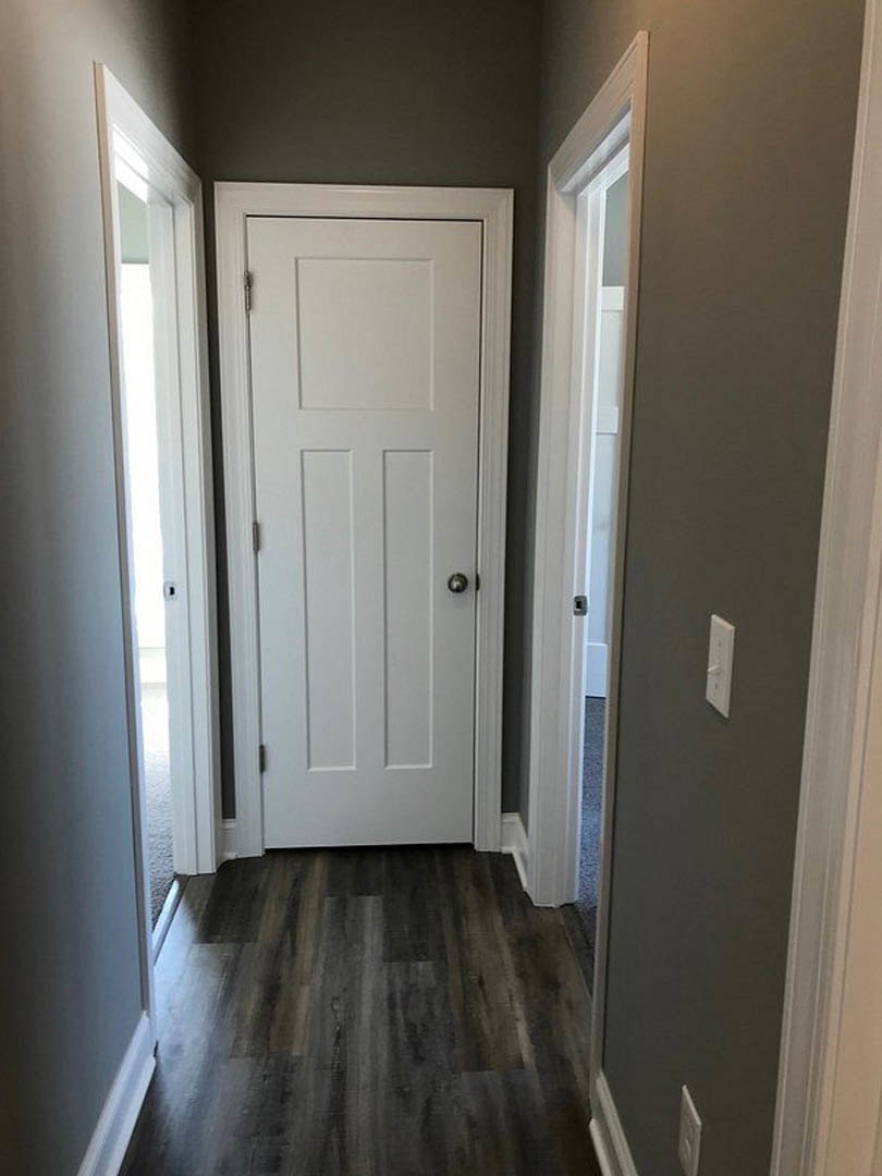 White door with silver knob and black trim in hallway with dark wood flooring and light switch on adjacent wall
