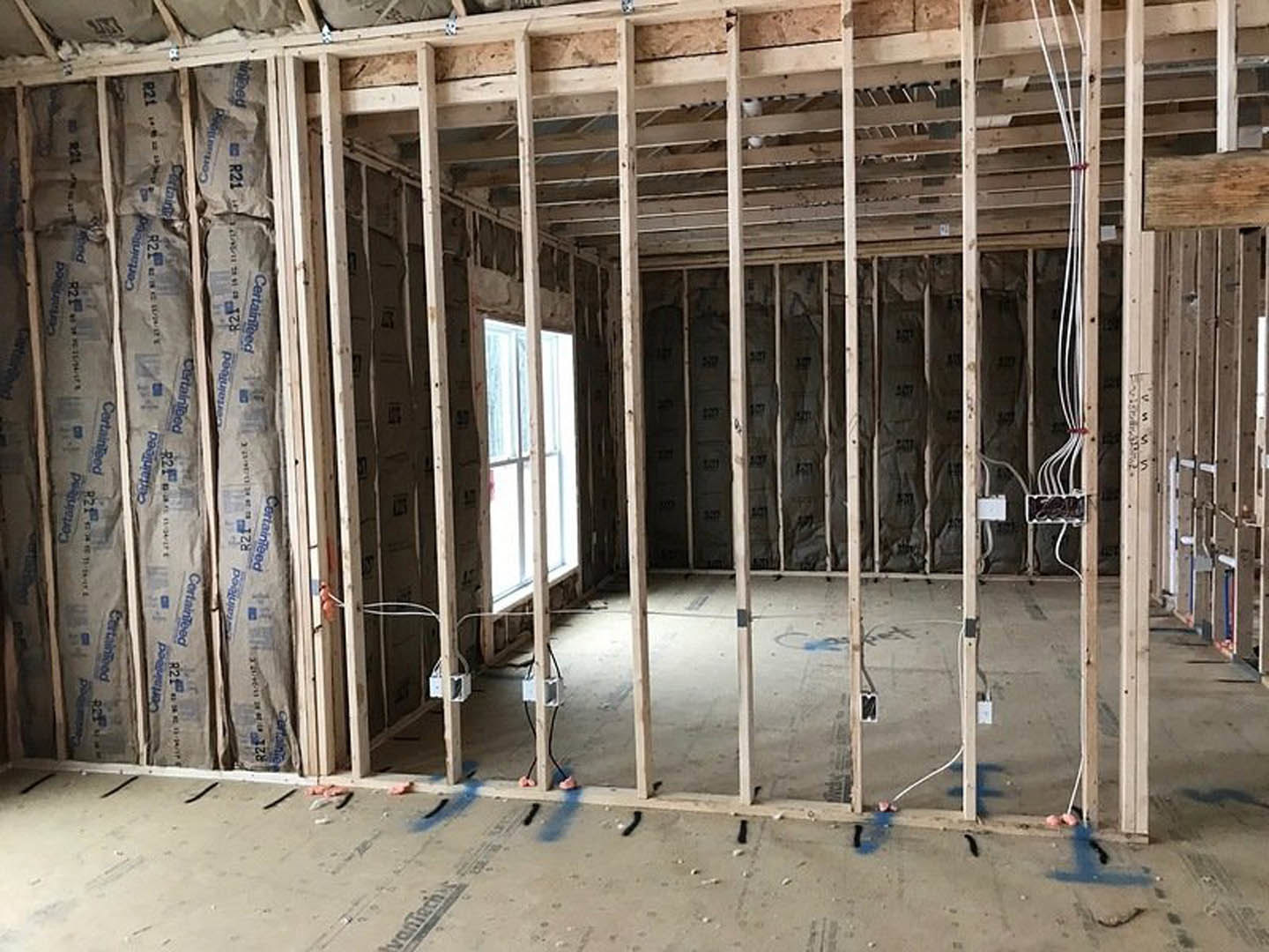 Wood-framed room under construction with exposed electrical wires, bags of cement on the floor, and sunlight streaming through a window