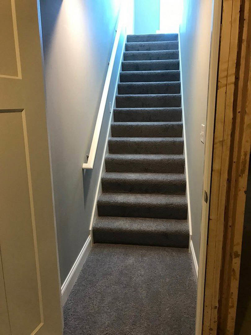 Carpeted staircase with wooden handrail, illuminated by overhead light, adjacent to painted wall and interior door