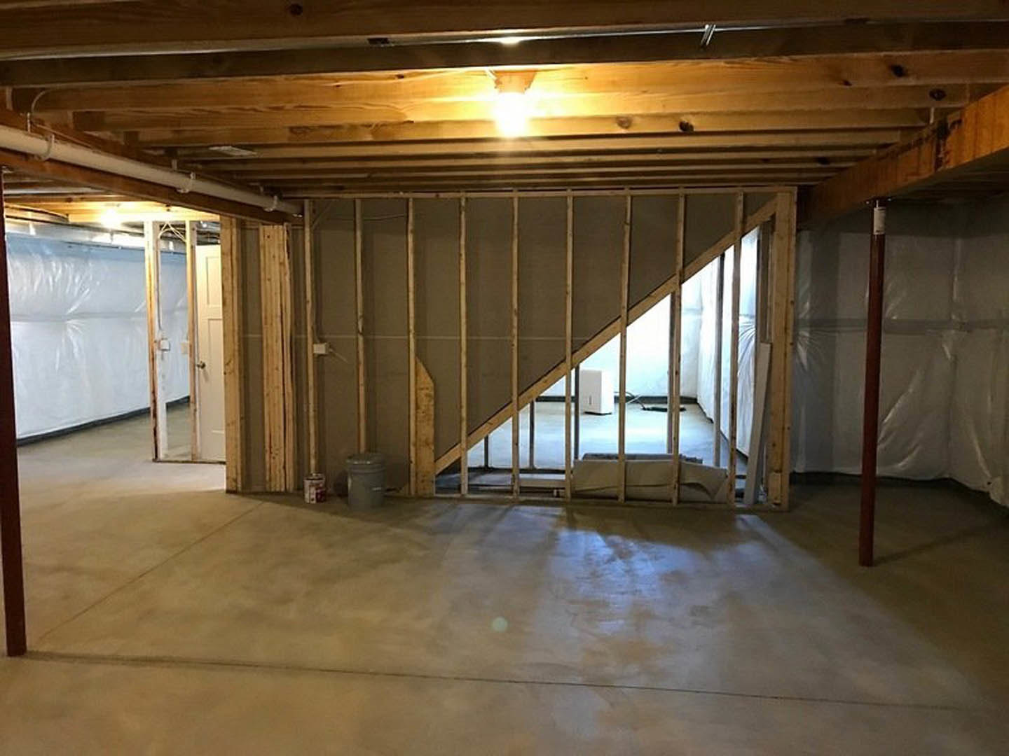 Unfinished room with exposed concrete floor, white plastic sheeting covering part of the wall, staircase partially visible, and ceiling illuminated by overhead light.
