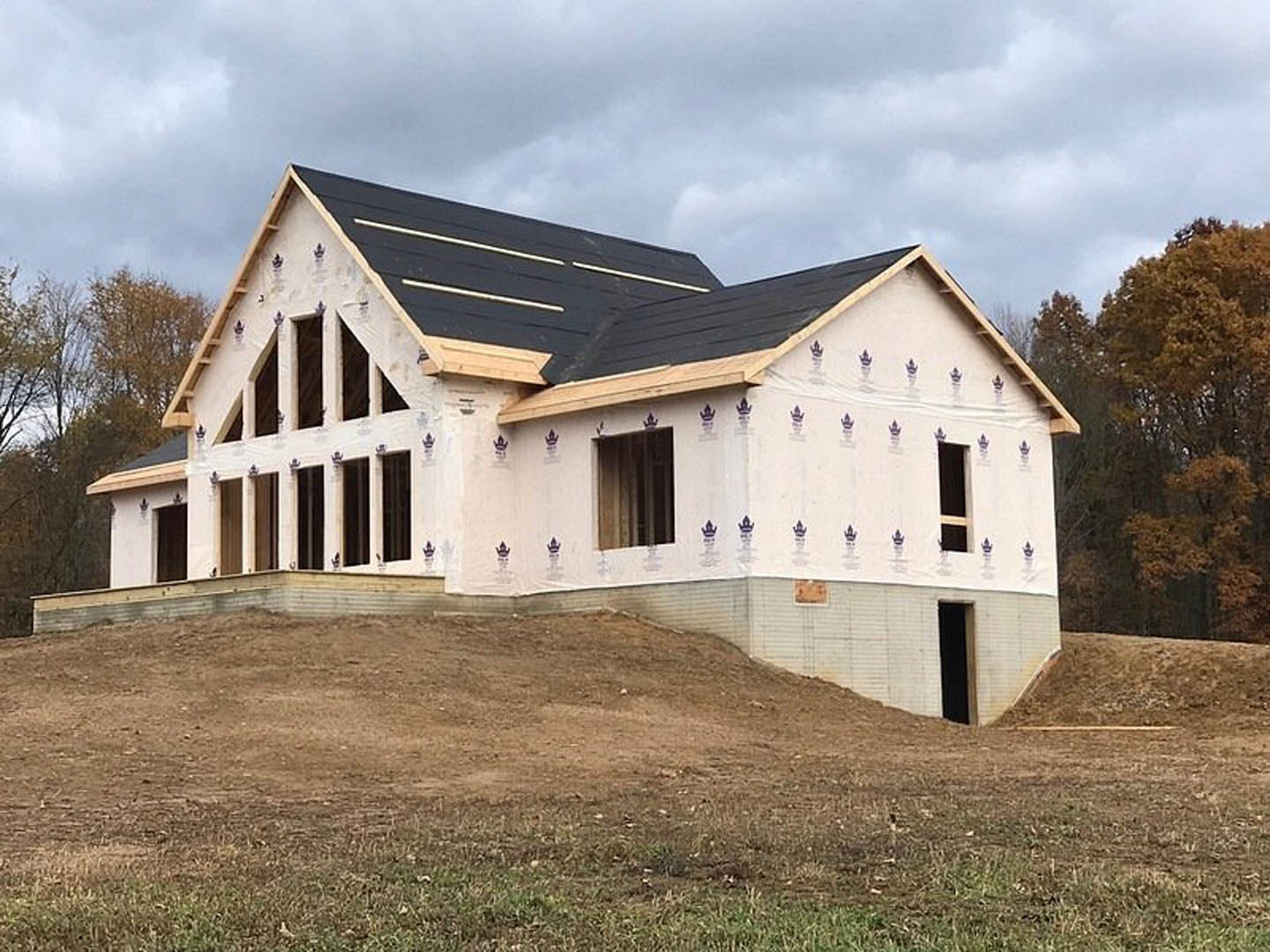 Wood-framed house under construction with exposed beams, dirt foundation, and white wall, surrounded by leafless trees and Robert Frost Farm visible in the background under a blue