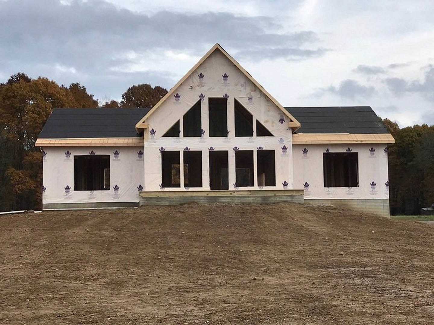 Two-story house under construction with exposed wooden framing, triangular roof, and dirt-filled yard; white windows installed, surrounded by trees and cloudy sky.