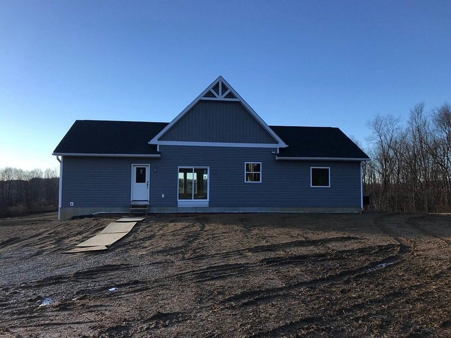 Modern cottage-style house with triangular roof, glass window, light siding, surrounded by trees, dirt field in foreground, broken wood planks scattered on ground