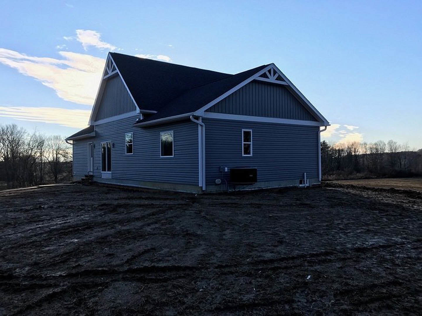 Partially built house with black shingle roof, exposed framing, white window insert, surrounded by dirt field with tire tracks, leafless trees in background
