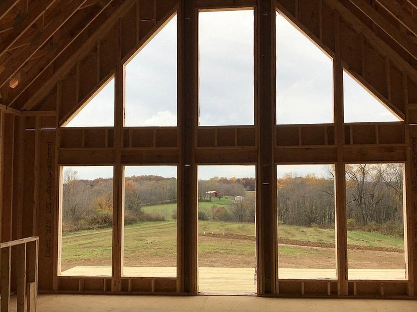 Floor-to-ceiling wood-framed window overlooking grassy field and distant trees, white cloudy sky visible, interior features wood paneling and beams