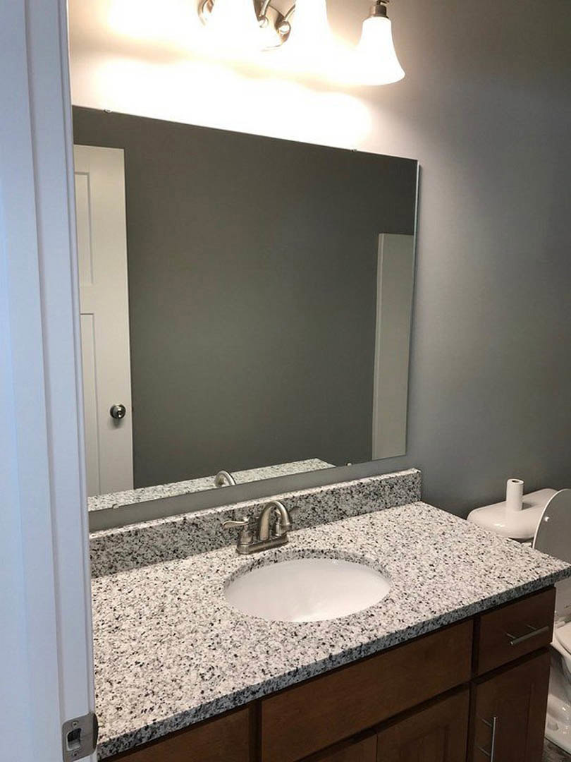 Modern bathroom with white tile walls, rectangular mirror above a stone countertop, chrome faucet and sink, white plate on the counter, toilet with toilet paper roll on tank, light