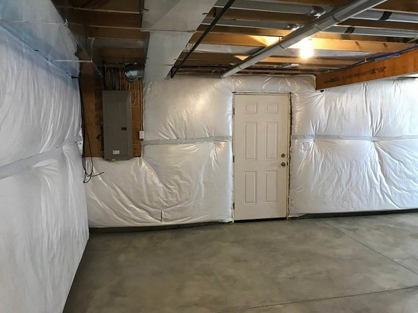 White door with silver handle set in a room with concrete floor, wall partially covered in translucent plastic sheeting, black square fixture visible on white wall.