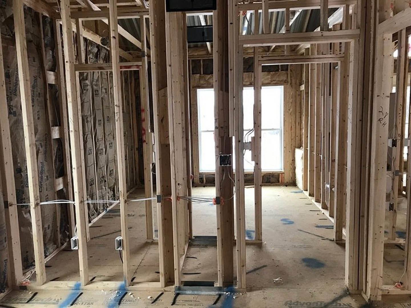 Unfinished room with exposed wood framing, white-framed square window, metal hardware, and insulation panels illuminated by natural light