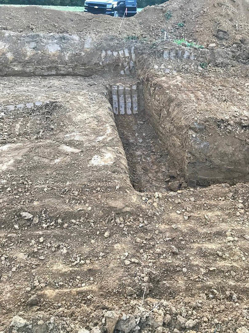 Deep trench dug into brown soil beside a parked vehicle, with exposed earth and scattered rocks visible along the edges.