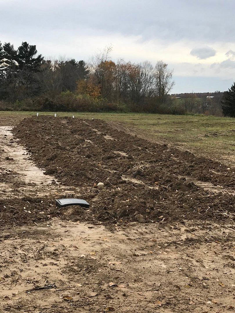 Dirt field with a television set placed on the ground, scattered green grass, dirt path, and cluster of leafy trees in the background under a partly cloudy sky
