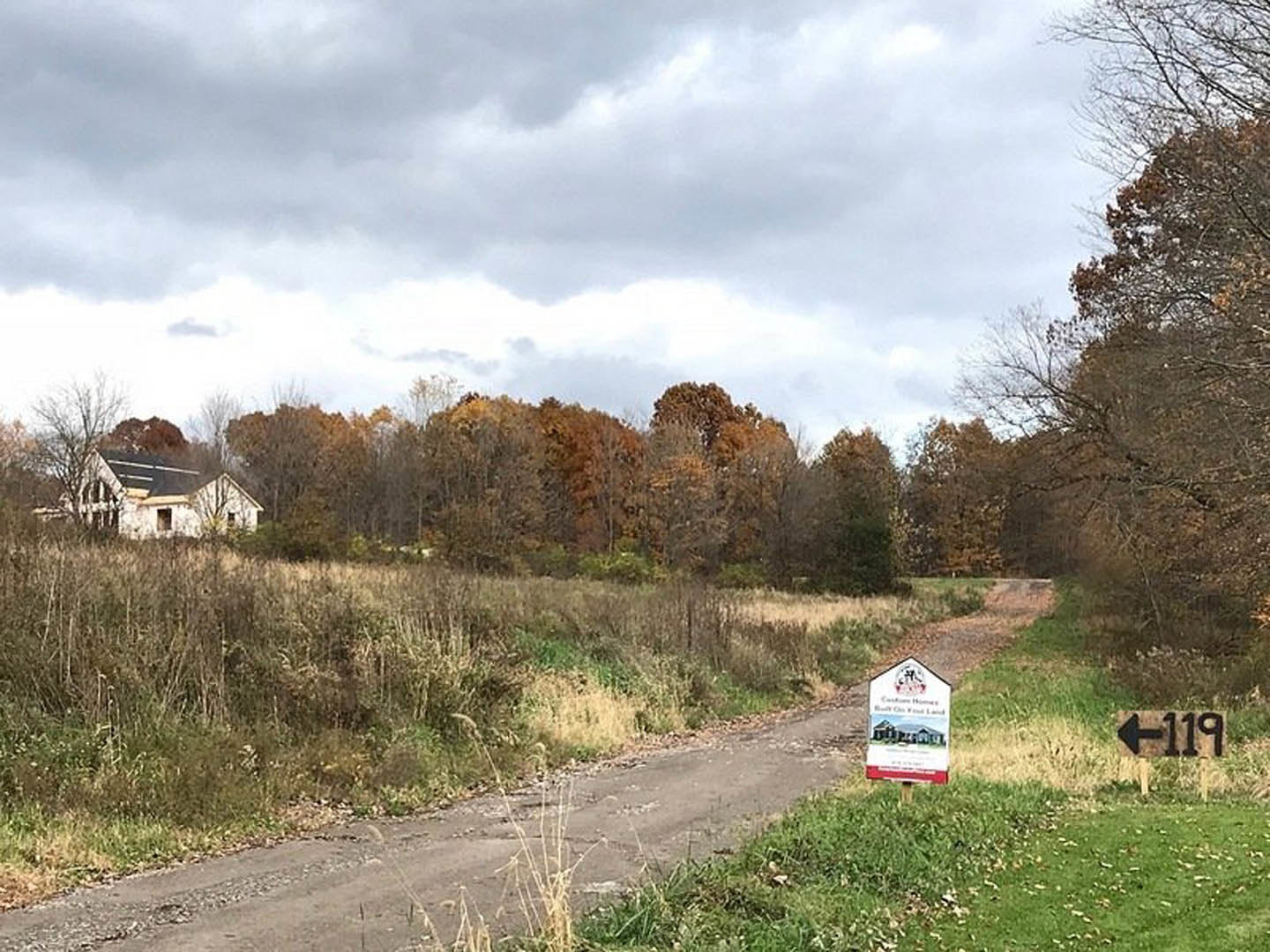 Black-roofed house beside a dirt road lined with bushes and plants, numbered sign with arrow in foreground, cloudy sky above autumn trees in rural setting