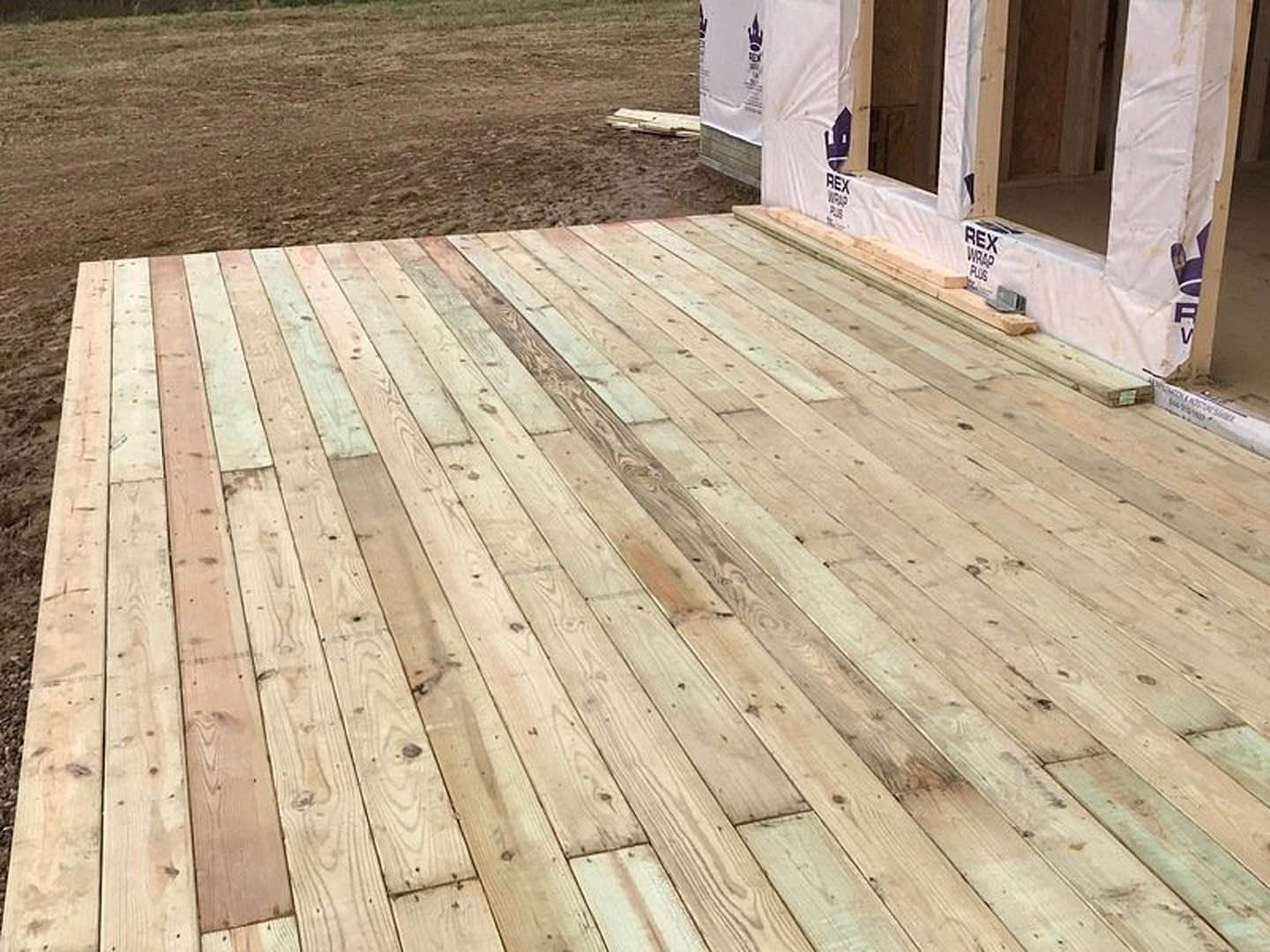 Wooden deck with unfinished house featuring exposed framing and white plastic barrier, surrounded by dirt field