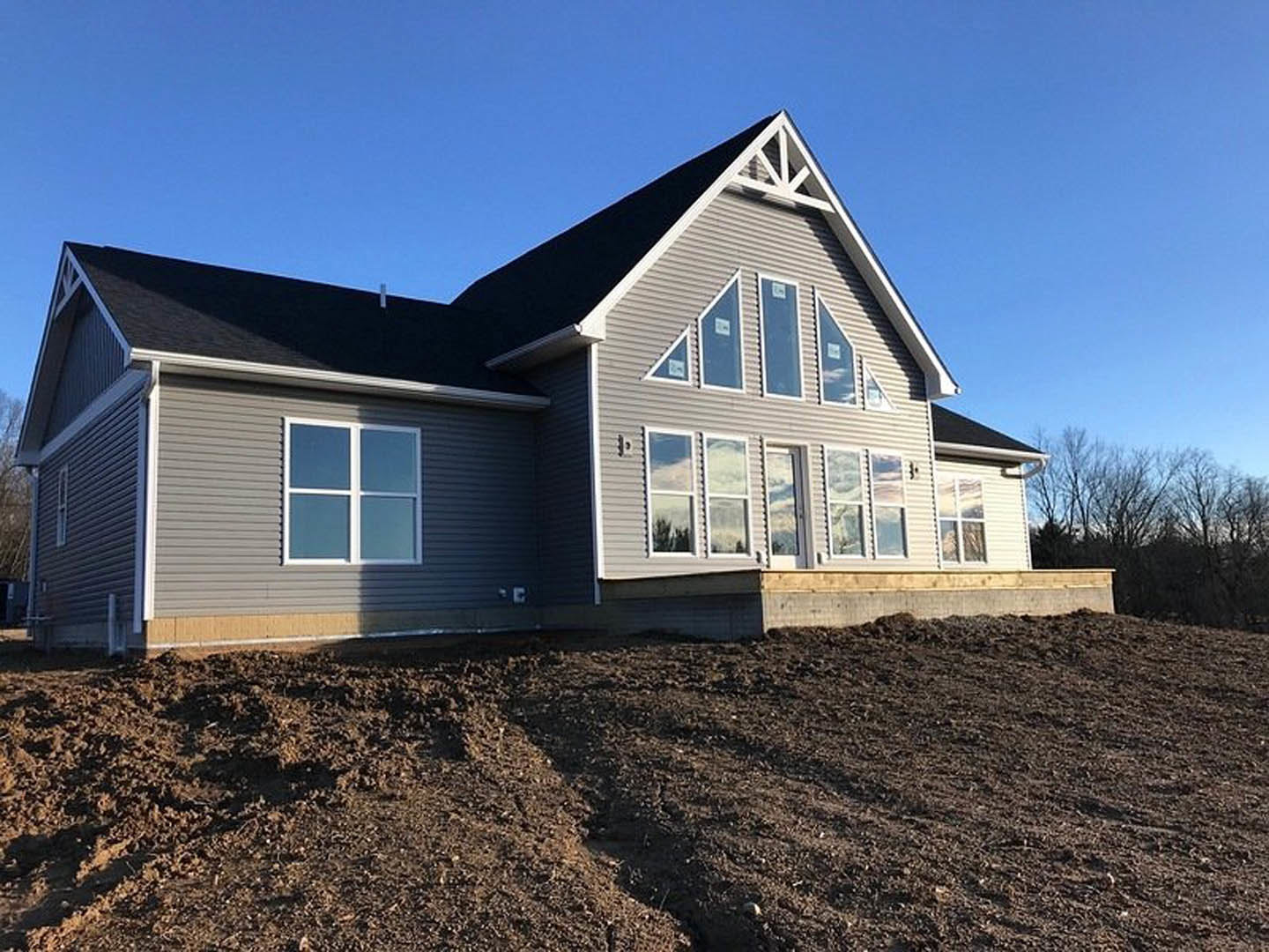 Modern house under construction with white-framed windows, light siding, and a large dirt field in the foreground under a clear blue sky