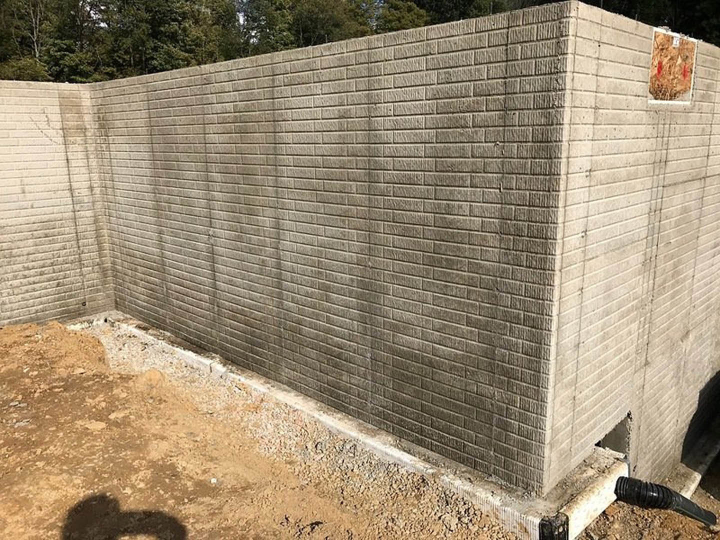 Concrete block wall with exposed pipe, plywood board nearby, black hose on wood surface, shadow of person cast on ground.