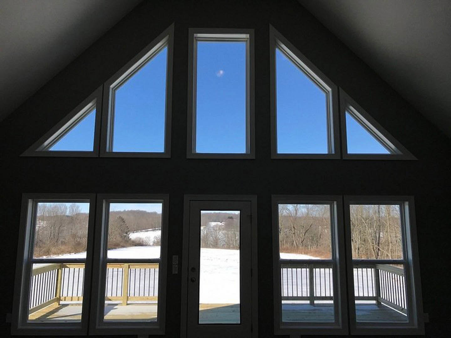 White custom home featuring a wooden deck and second-story balcony, glass-paneled door opening to a snowy landscape, moon visible in the sky through windows.