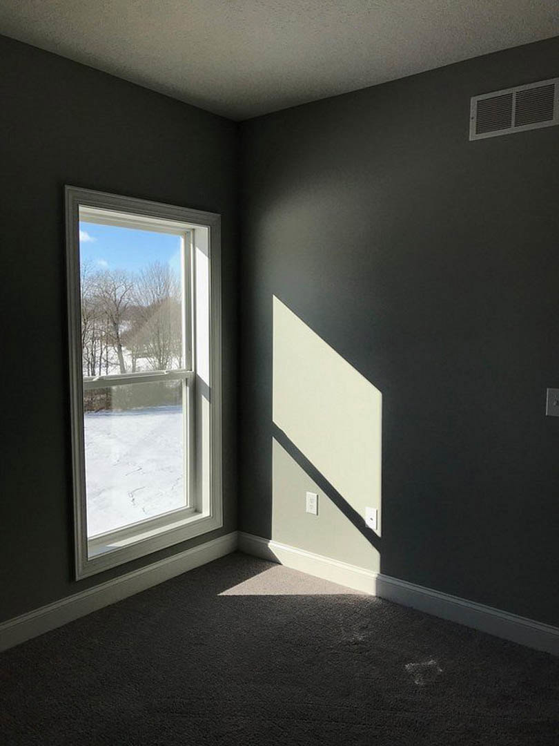 Large window overlooking snowy trees, white plaster walls, dark carpeted floor, rectangular vent on wall, natural daylight filling the room